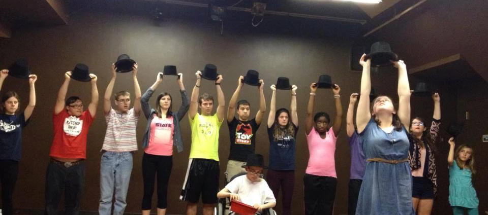 Students from the Harmony musical theatre workshop raise top hats over their heads during a performance.